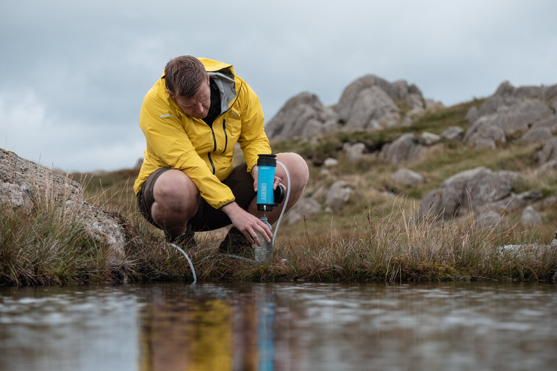 Load image into Gallery viewer, Person in a yellow jacket collecting water from a stream in a rocky landscape

