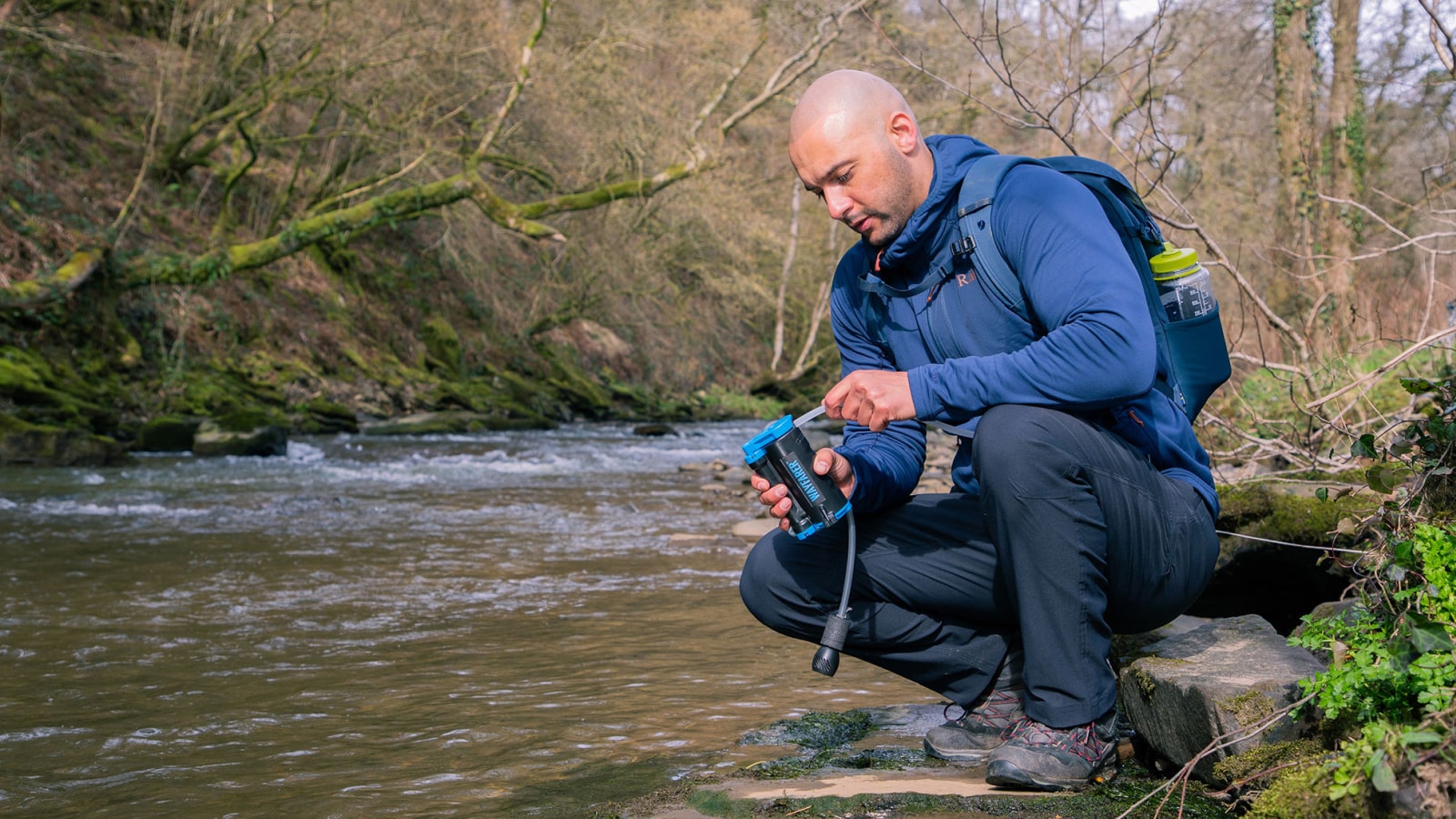 Man with a backpack by a river, using a water filter device.